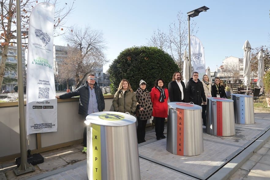 Ανθρώποι σε χώρο ανακύκλωσης στη Λάρισα με κάδους και ενημερωτικές πινακίδες.