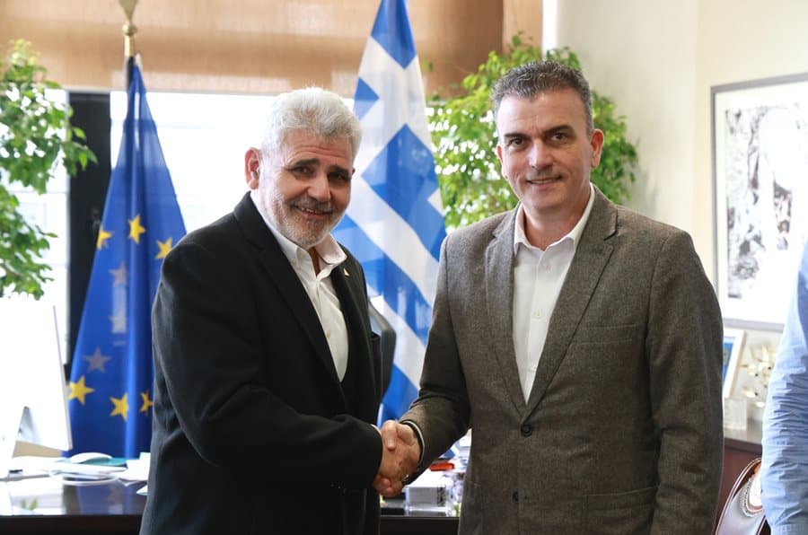 Δύο άνδρες σε χειραψία με σημαίες Ελλάδας και Ευρωπαϊκής Ένωσης στο φόντο.