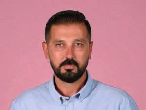 Ανδρας με σκούρα μαλλιά και μούσι, φοράει γαλάζιο πουκάμισο, μπροστά σε ροζ φόντο.