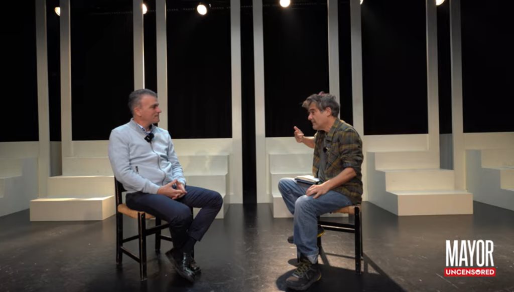 Άνδρες σε συζήτηση σε σκηνή, με φόντο ασπρόμαυρα παράθυρα, συζητούν ανοιχτά.